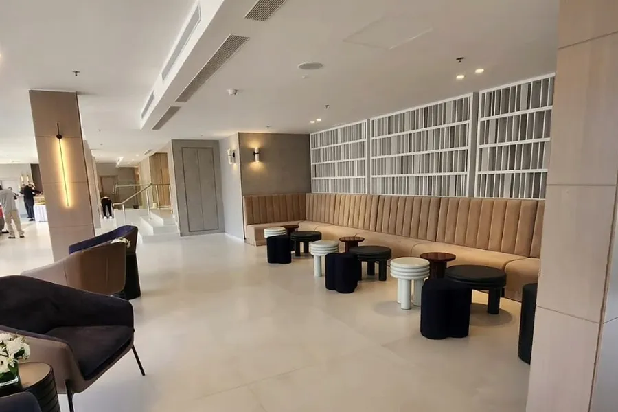לובי מלון אסטרל וילג' אחרי השיפוץ: קיר ספרייה לבן, ספסל ישיבה מרופד, שרפרפים עגולים ותקרת גבס עם תאורה