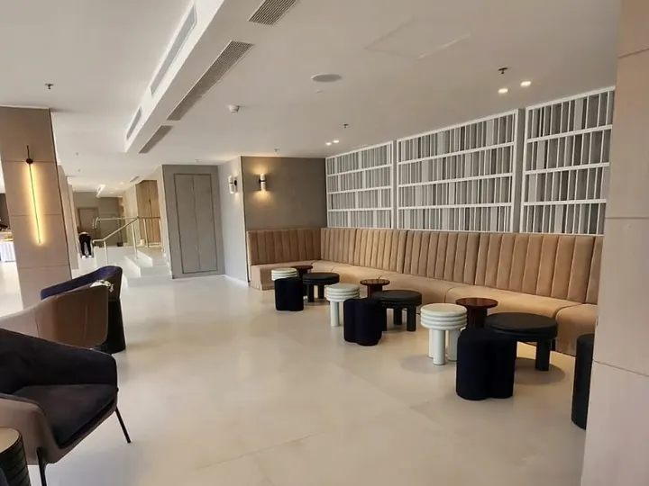 לובי מלון אסטרל וילג' אחרי השיפוץ: קיר ספרייה לבן, ספסל ישיבה מרופד, שרפרפים עגולים ותקרת גבס עם תאורה